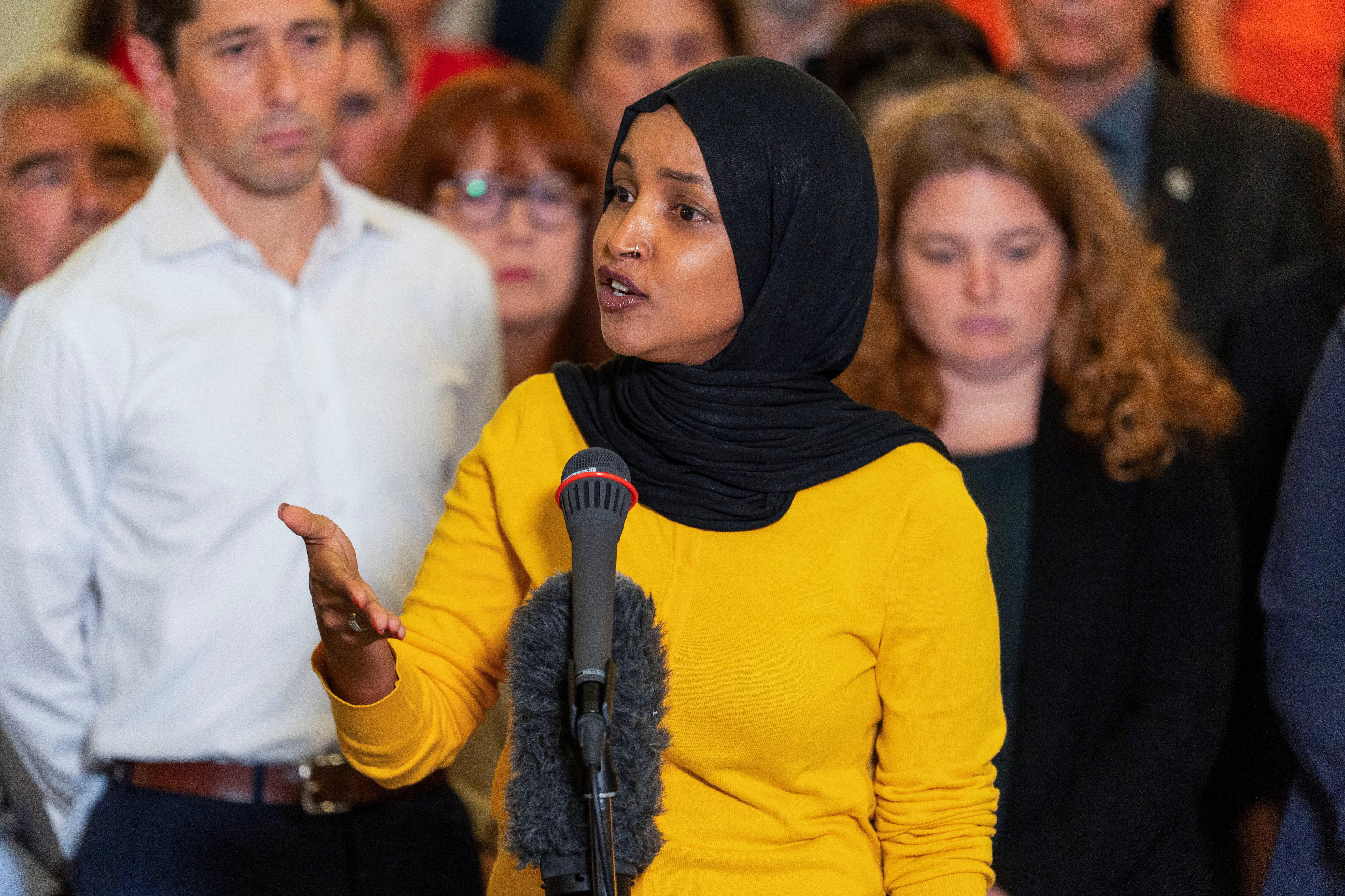 Ilhan Omar speaks animatedly, wearing a yellow dress and a black headscarf.