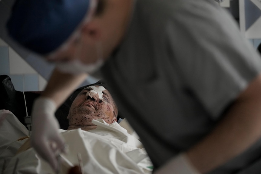 A medic attends to a man lying in a bed with bandages and scars on his face.