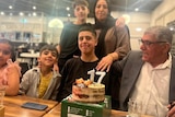 A family poses for a photograph at a 17th birthday party. Parents have their arms around their two sons, and two grandchildren