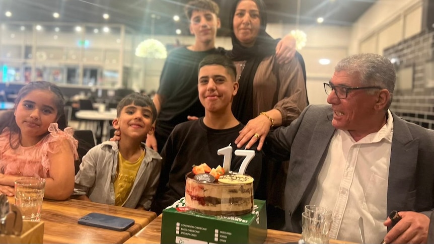 A family poses for a photograph at a 17th birthday party. Parents have their arms around their two sons, and two grandchildren