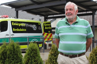 A man with white hair in a green and white shirt stands in front of an ambulance that's parked underneath a shelter.