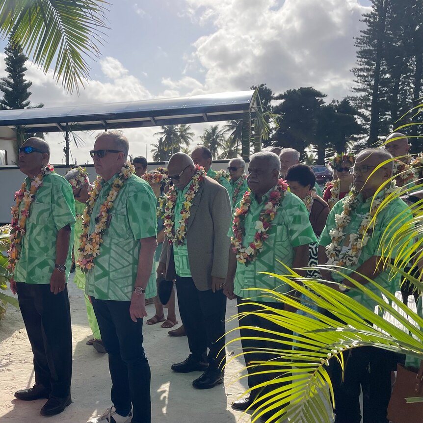 Anthony Albanese and other Pacific leaders wear green shirts and flower necklaces as they stand in a group in sunlight.