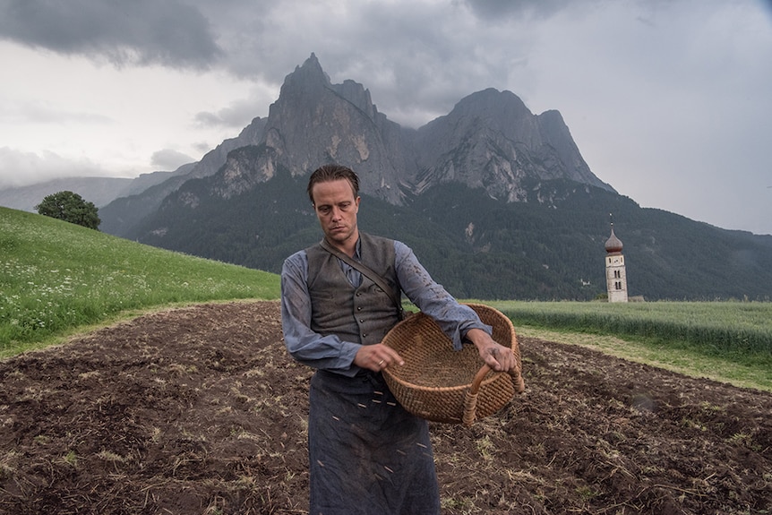 A man in peasant farmer attire with serious expression looks down and throws seeds on land in countryside on an overcast day.
