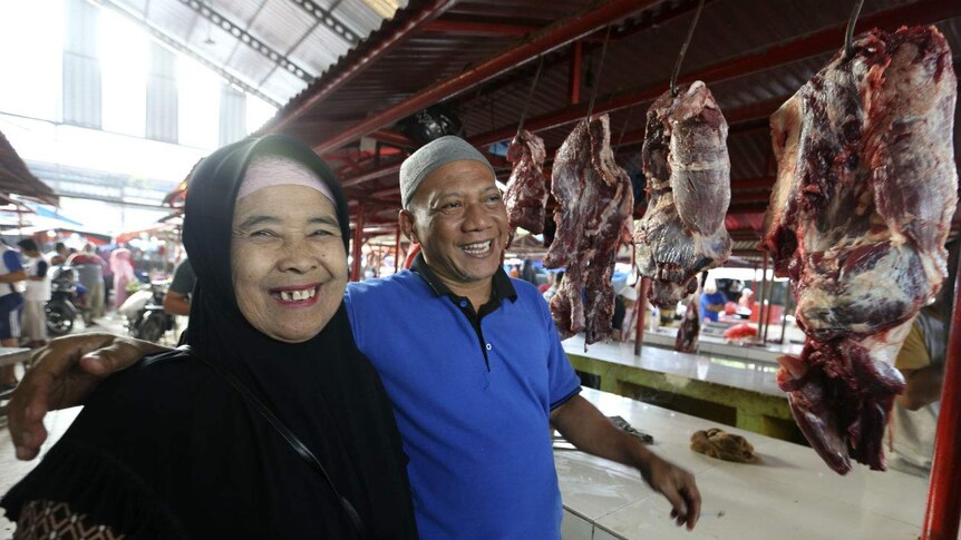 A butcher in an Islamic cap smiles as he puts his arm around a female customer in a headscarf at a butchers stall at markets.
