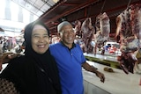 A butcher in an Islamic cap smiles as he puts his arm around a female customer in a headscarf at a butchers stall at markets.