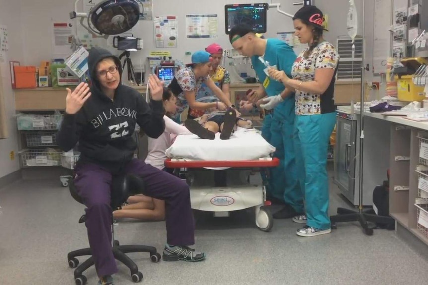 A doctor wearing a hoodie over her scrubs dances in front of a hospital bed with young people wearing scrubs in attendance.