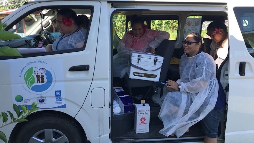 The side of a white van has its sliding door open as medical staff in protective clothing carry measles vaccines.