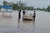 Three young men wearing black standing next to a white engine boat on a flooded road next to blue road sign.