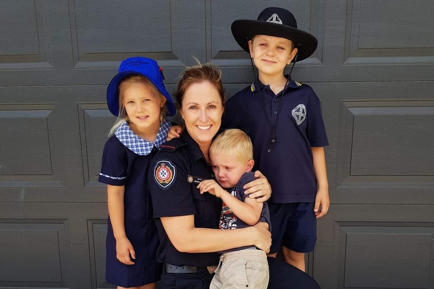 A mum dressed in a firefighter uniform kneels down and hugs three young kids.