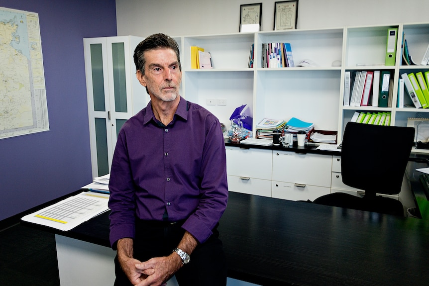 A man in a purple shirt sits on an office desk and looks off to the distance.