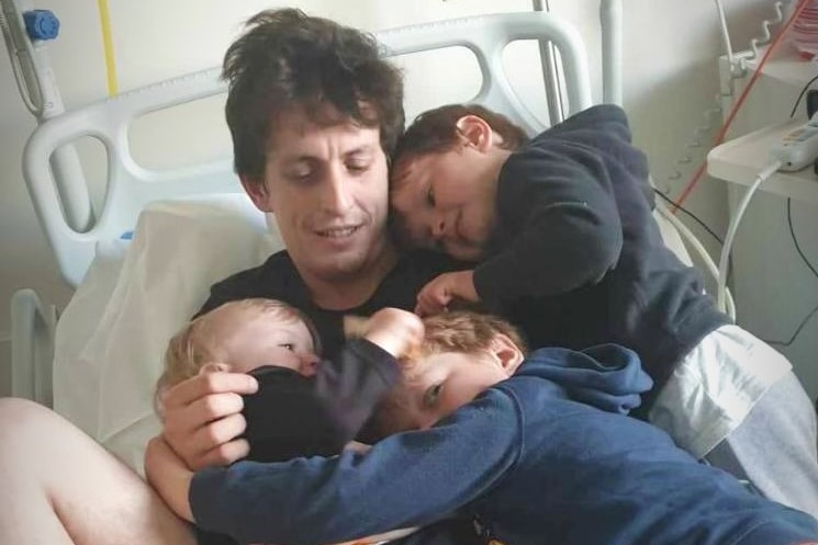 A man with brown hair lies in a hospital bed with three small children cuddling him.
