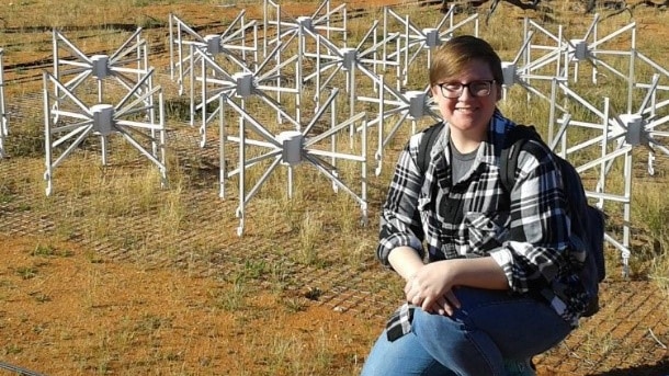A woman squatting in the red dirt in front of a series of white sci-fi looking radio telescopes.