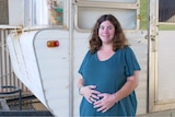 A pregnant woman stands in front of a caravan where a cat can be seen looking at her through a window.
