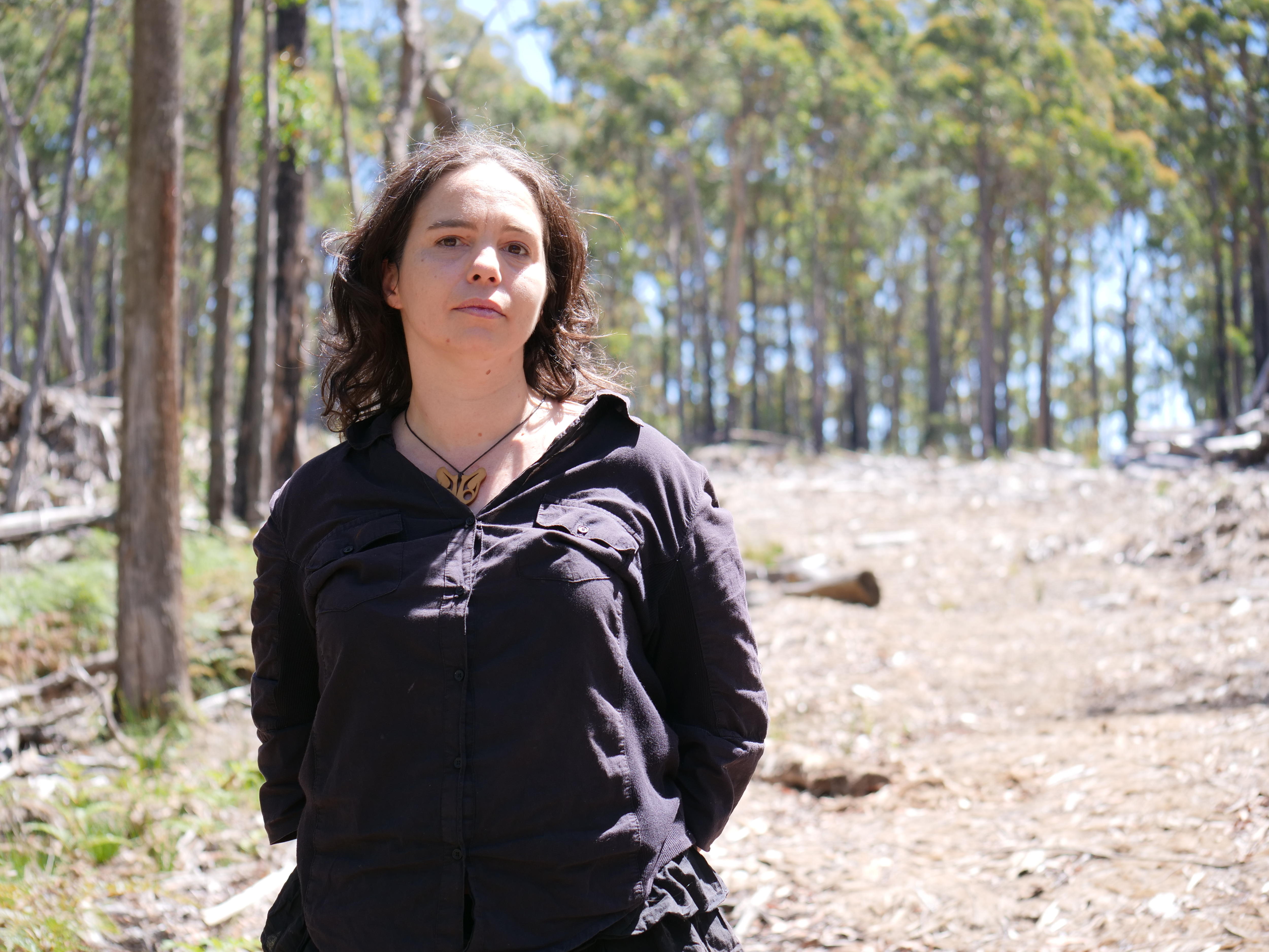 A woman stands in front of a cleared patch of forest. 