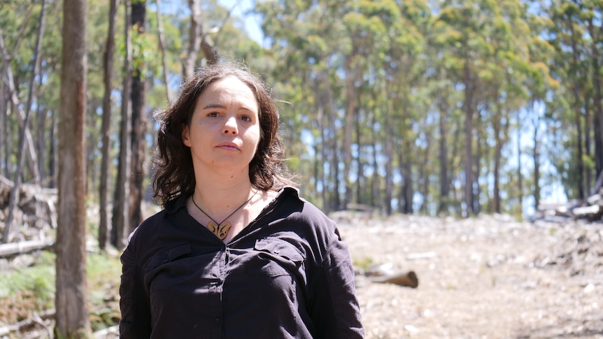 A woman stands in front of a cleared patch of forest. 