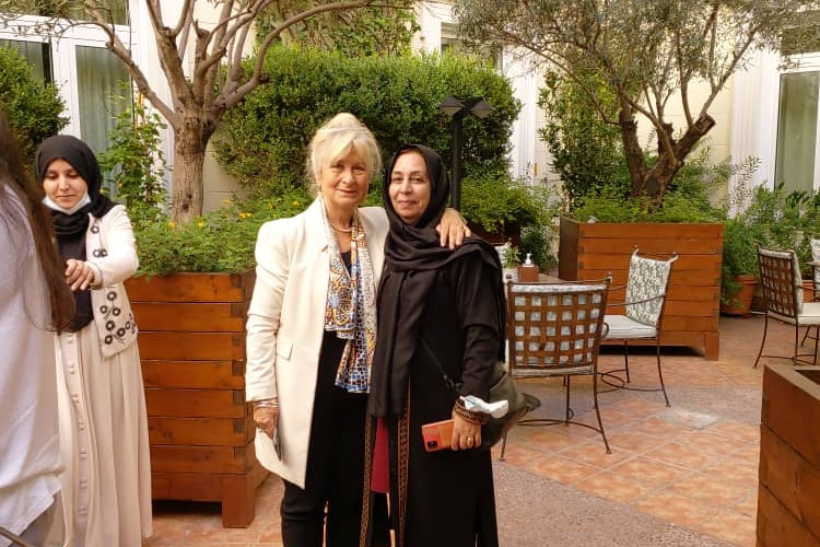 A woman with white hair and wearing a jacket puts her arm around a woman wearing a hijab and black jacket