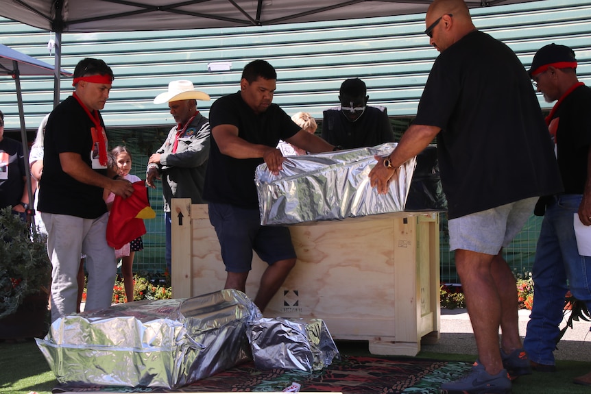 Two men taking two large boxes out of a larger wooden box and placing them on the ground.