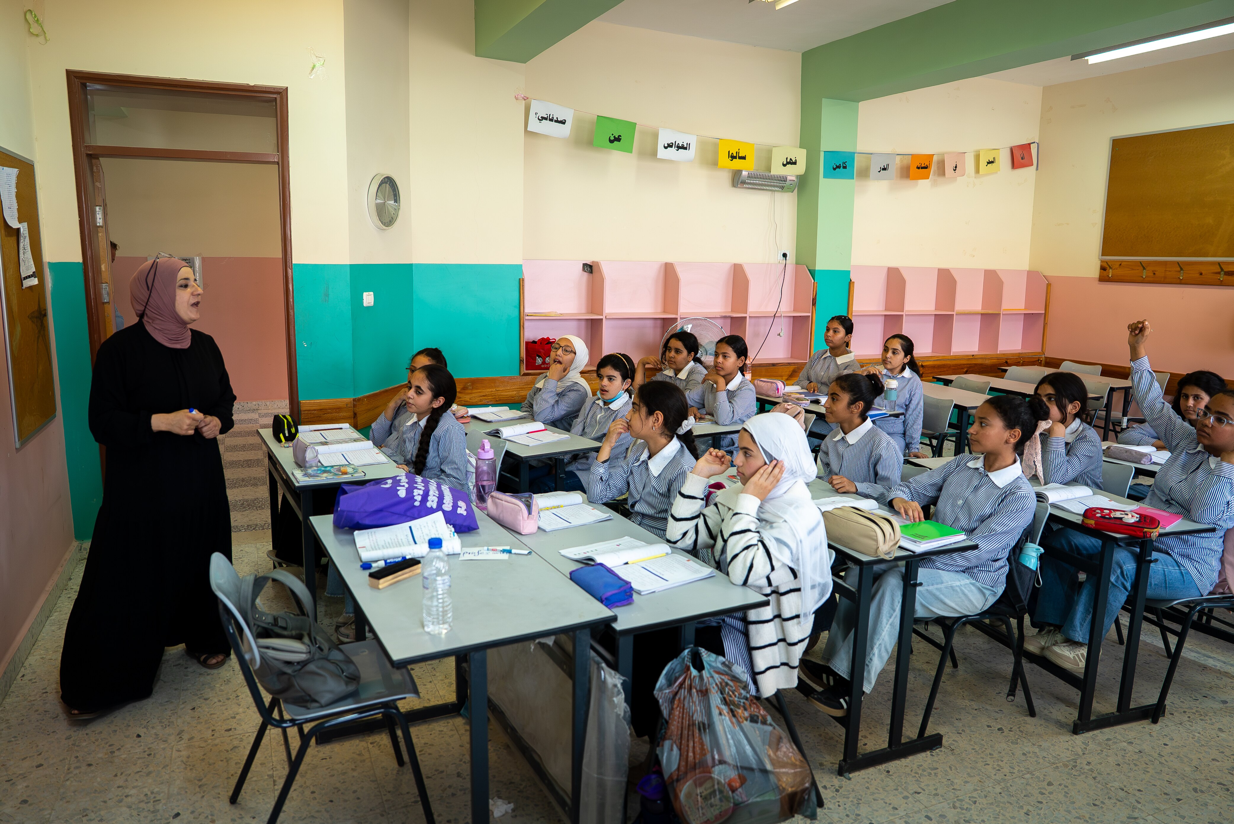 A group of students sit at their desks listening to a teacher at the front of the classroom.
