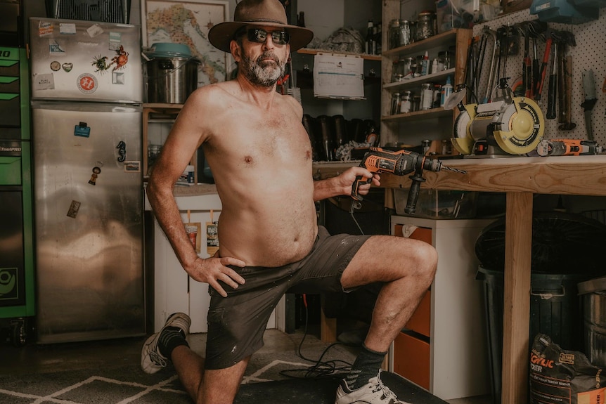 Man kneeling on one knee at a work bench with hat and sunnies on, holding a drill while not smiling