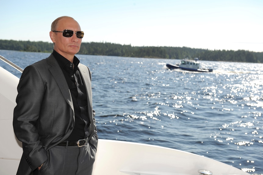 Vladimir Putin in sunglasses, a dark suit and black shirt stands on a boat looking at the water