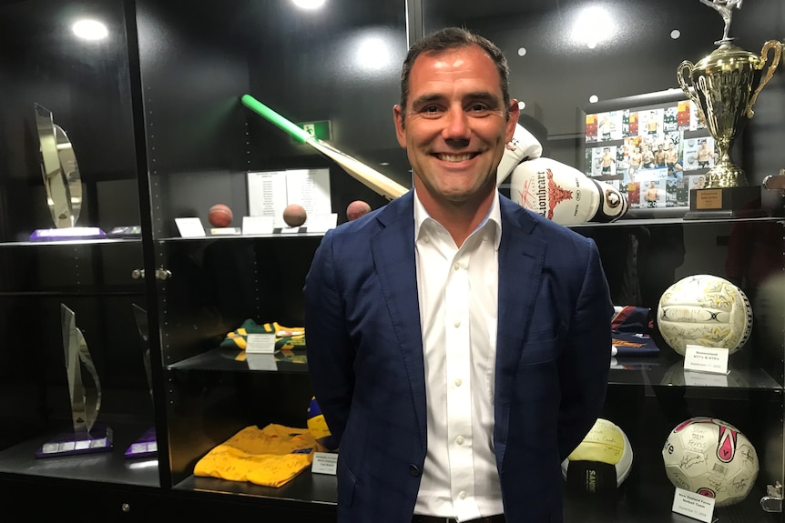 An athletic middle-aged man in navy blue blazer and white shirt smiles at camera in front of sports trophy cabinet