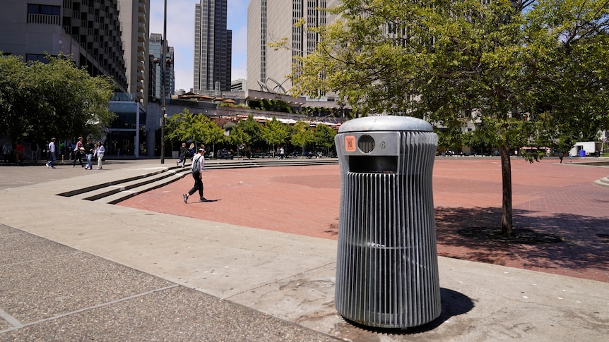 A short, round, metal-slatted garbage bin sits in an open concrete space in front of a tree-lined cityscape.