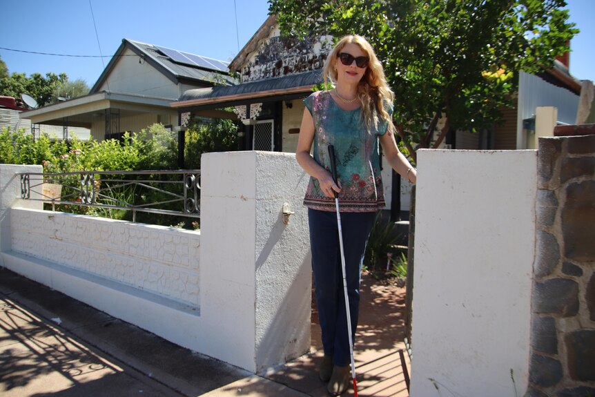 A vision impaired woman in glasses holding a cane and standing by a white concrete fence