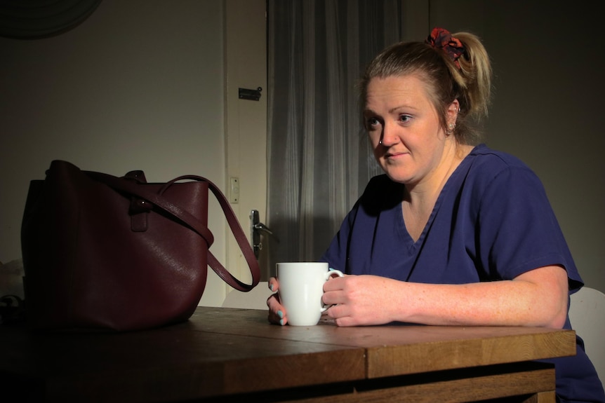 Kim Gallaher, dressed in blue scrubs, sits at a table with a mug.