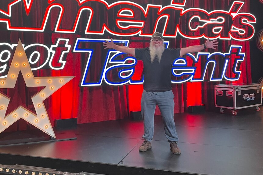 A bearded man in a cap with glasses standing in front of a neon America's Got Talent sign