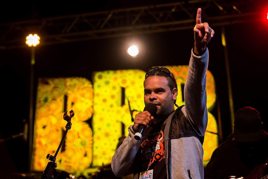 A singer stands on a stage with a microphone in front of large sign that reads 'BBB'.