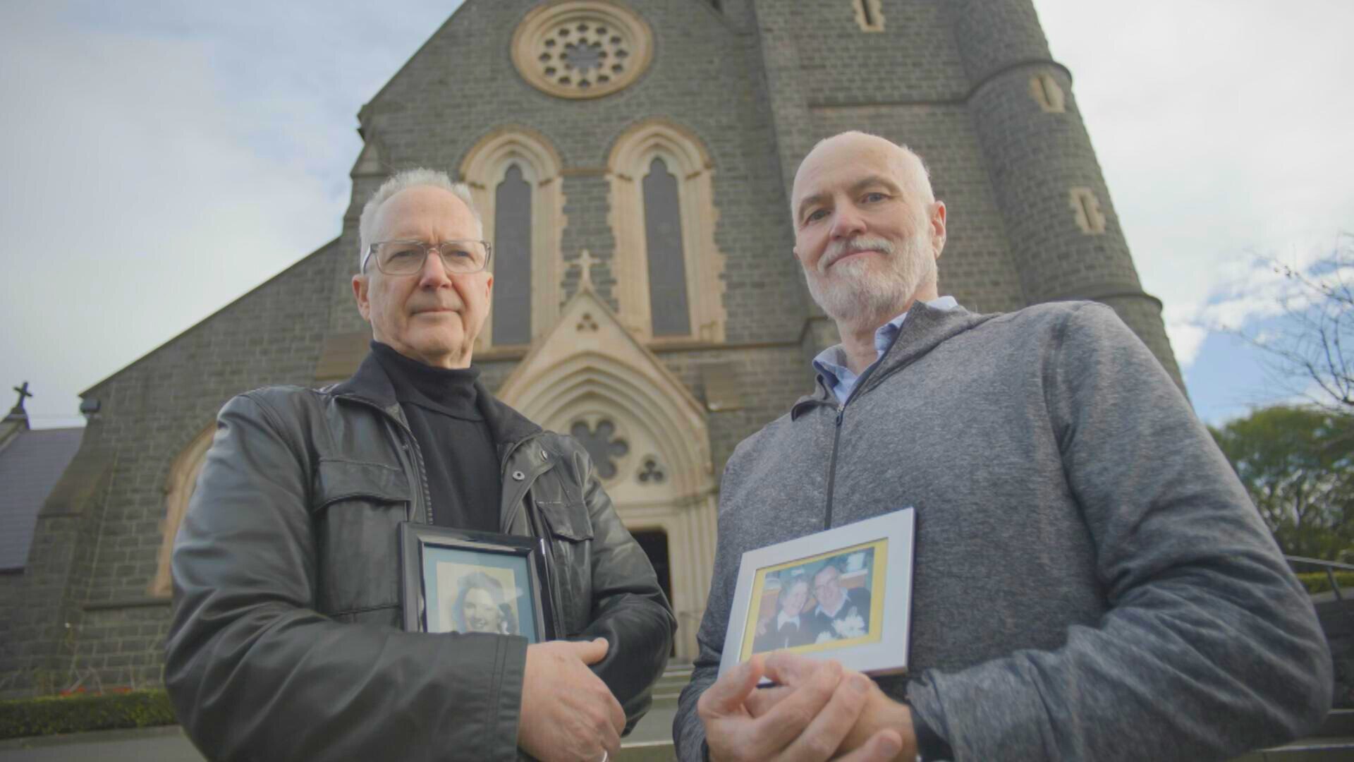 Two men standing in front of a church holding photos in frames of their parents