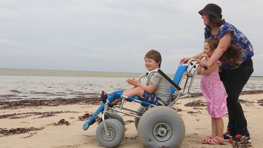 Boy in beach wheelchair on right being pushed by mum and sister standing, all on beach