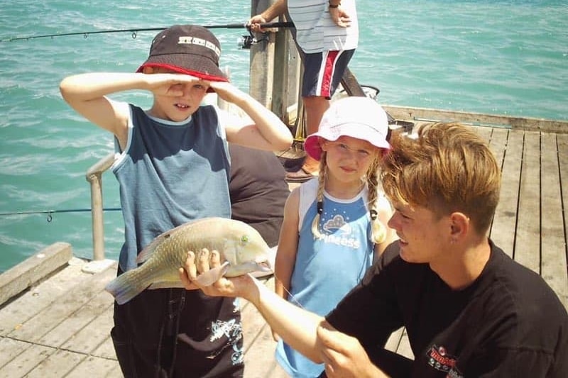 a young man kneels down holding a fish next two two young children 