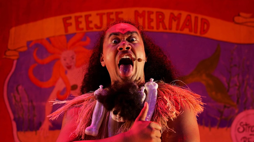 Red-lit with banner backdrop reading "Feejee Mermaid", with performer centre staring at camera pull fierce face with tongue out.
