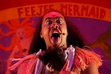 Red-lit with banner backdrop reading "Feejee Mermaid", with performer centre staring at camera pull fierce face with tongue out.