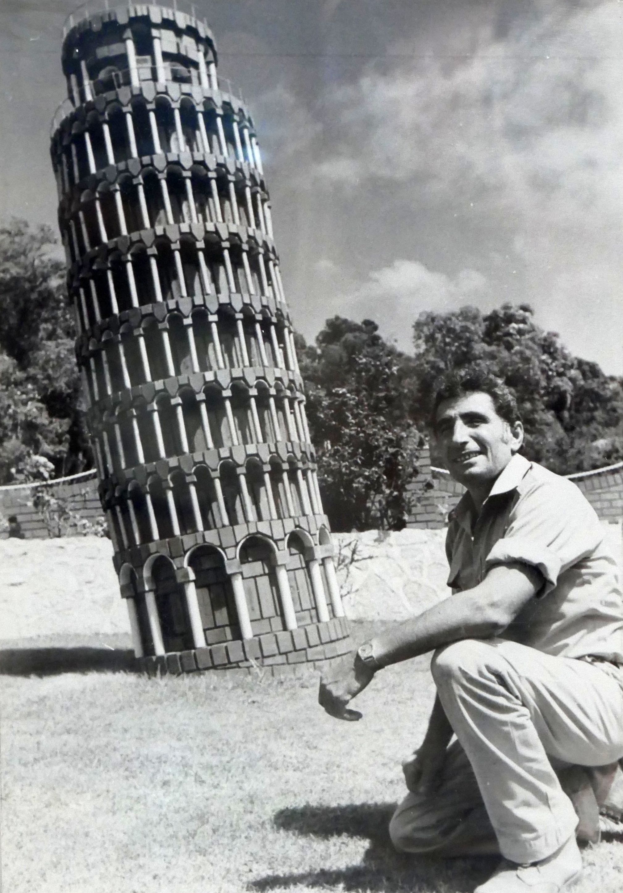 A black and white photo of a man next to a replica of the Leaning Tower of Pisa.