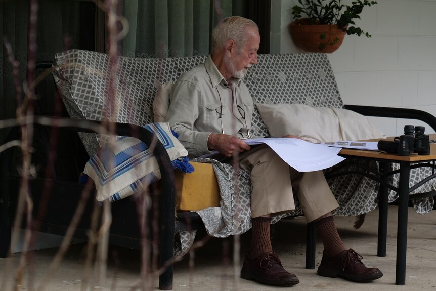 An older man sits on a lounge seat outdoors, looking through old photos.
