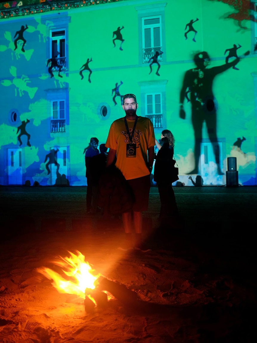 Myles Walsh stands in front of a two storey building partly lit by the images projected onto the building behind him.
