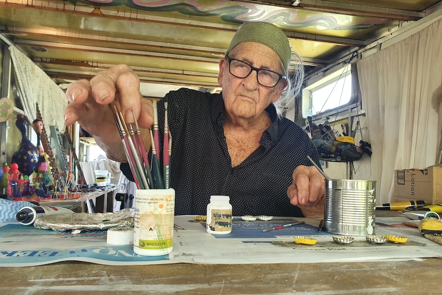 An old man, wearing a green headband, glasses, a t-shirt with blue dots, chooses a paintbrush while sitting at a desk.