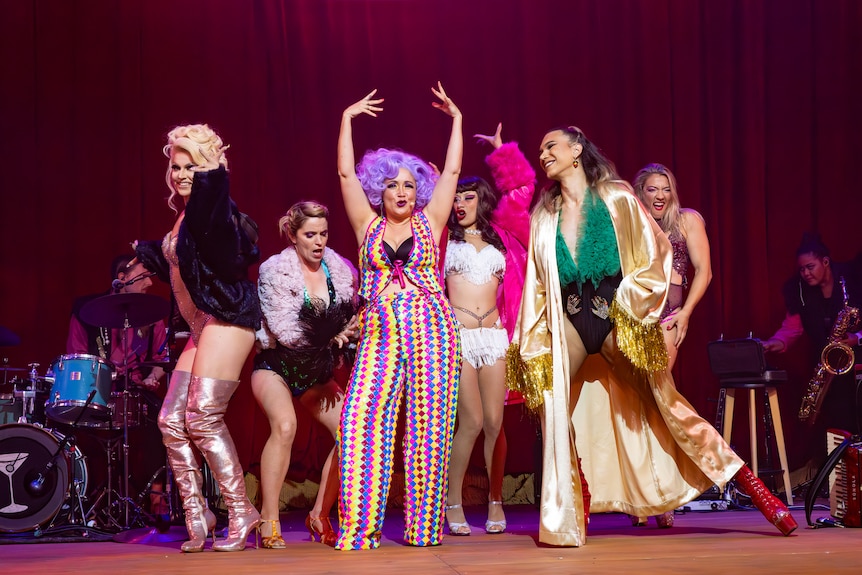 A group of femmes in colourful outfits and wigs, dance together in a burlesque-style onstage.