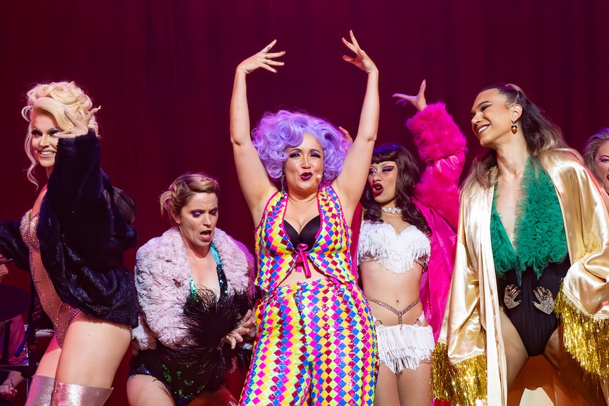 A group of femmes in colourful outfits and wigs, dance together in a burlesque-style onstage.