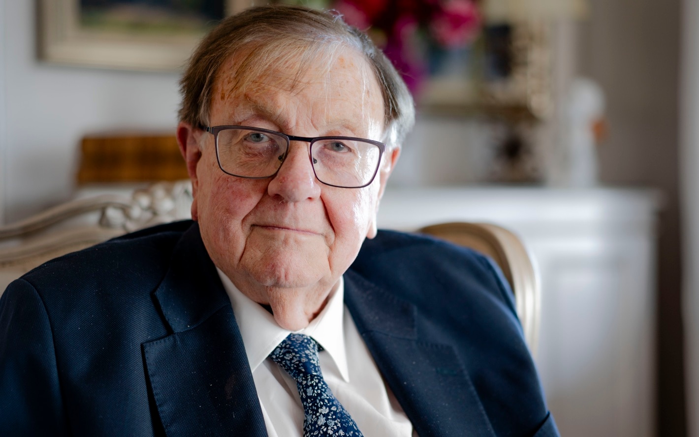 Anthony de terno, óculos, sorri severamente, é mais velho, tem cabelo cada vez mais curto, senta em uma linda cadeira ornamentada e pinta borrada atrás.