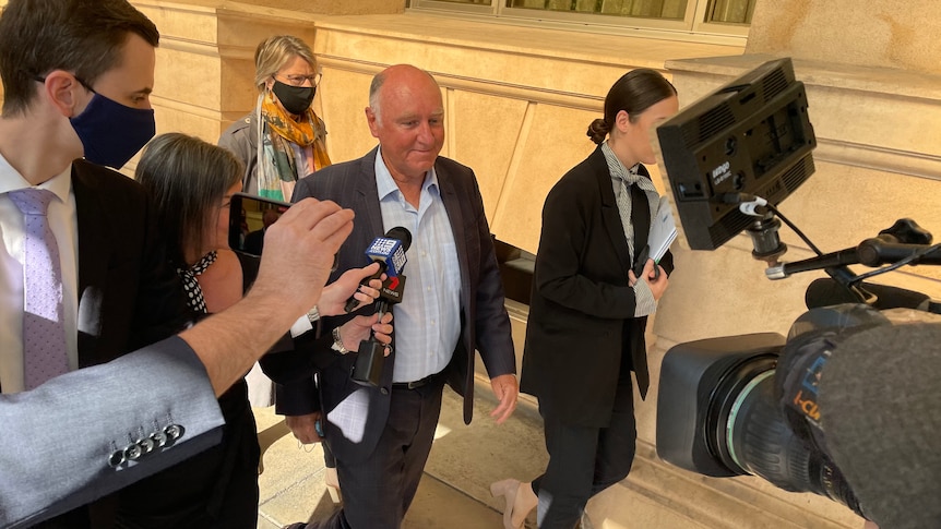 A bald man wearing a suit with no tie followed by journalists and a TV camera