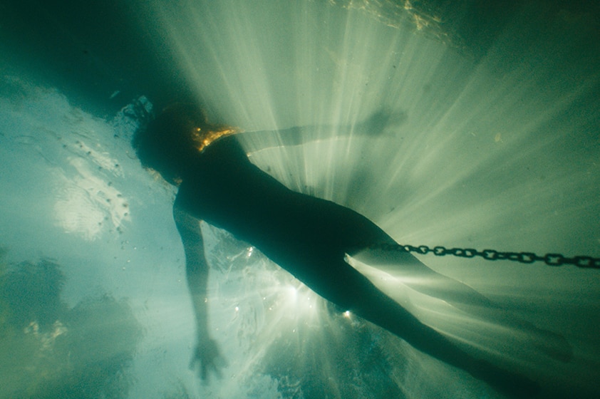 Silhouette of lone person floating face up in water connected to chain, rays of light shine through the top of the water.