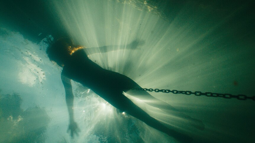 Silhouette of lone person floating face up in water connected to chain, rays of light shine through the top of the water.