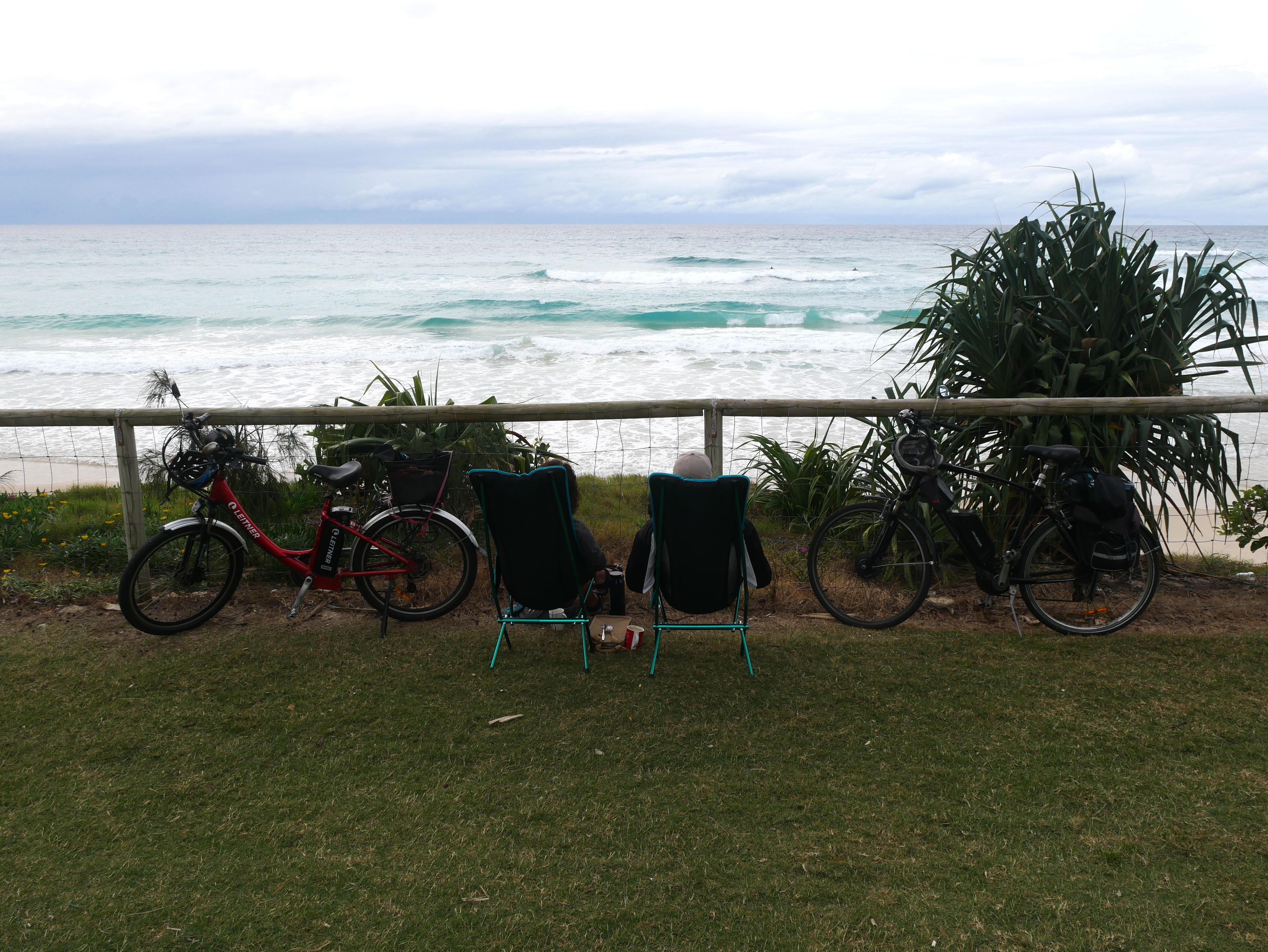 couple sitting in chairs looking out onto the ocean with their bikes leaning against the fence