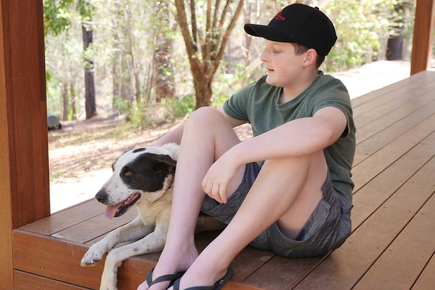 A young boy sits on a wooden deck with his arm around his black and white dog.