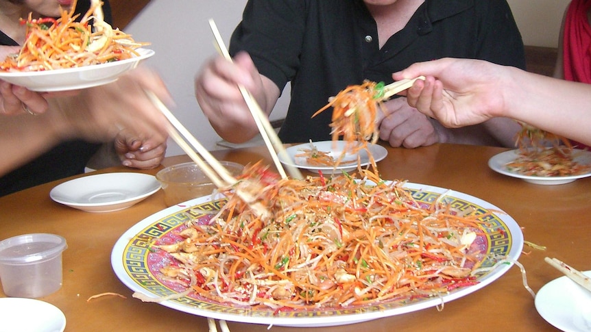 Three hands holding chopsticks dig in to a brightly coloured Yee sang noodle dish on a plate.