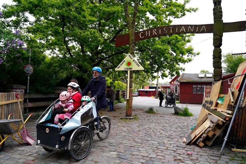 You view a young family in a box tricycle over a cobblestone laneway, with a sign behind them reading 'Christiania'.