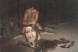 Indigenous man sits on crocodile holding a knife to it. The crocodile jaws are wedged open with a piece of wood.